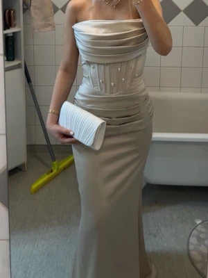 Beige långklänning med pärldetaljer - Elegant beige långklänning med axelbandslös design och figurnära passform. Klänningen har en vacker korsettdel med pärldetaljer och draperad överdel. Perfekt för speciella tillfällen.