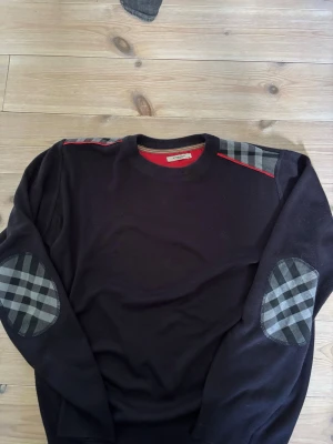 Svart tröja med rutiga detaljer Burberry - Svart tröja från Burberry med rutiga lappar på armbågar och axlar. Rund halsringning med röd insida och diskret broderad logga på bröstet. Klassisk och stilren design med snygga kontrastdetaljer.