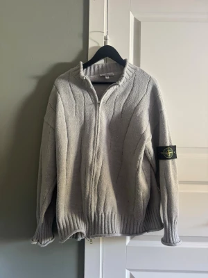 Grå ziptröja från Stone Island - Säljer en grå stickad ziptröja från Stone Island med ribbade muddar och krage. Tröjan har dragkedja framtill och den klassiska Stone Island-loggan på ärmen. Perfekt för lager-på-lager och har en avslappnad passform.
