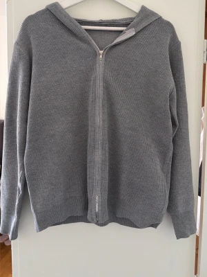 Grå stickad hoodie med dragkedja - Säljer en stilren grå stickad hoodie med dragkedja framtill. Hoodien har långa ärmar, ribbad struktur och en klassisk huva. Perfekt för en avslappnad och trendig look. Materialet känns mjukt och bekvämt mot huden.