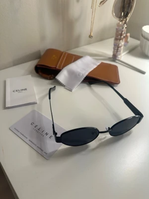 Svarta ovala solglasögon från Celine - Snygga svarta solglasögon från Celine med smal, oval form och minimalistisk design. Metallbågar och mörka glas ger en cool vibe. Kommer med originalfodral, putsduk och certifikat. Perfekta för dig som vill ha en stilren accessoar.