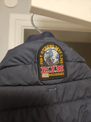Mörkblå dunväst från Parajumpers - Snygg mörkblå dunväst från Parajumpers med emblem på ryggen och hög krage. Västen har två fickor framtill med dragkedja. Strl. L 