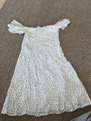 Vit kortklänning med små blommor - Söt vit kortklänning med blå blommor och smockad överdel. Klänningen har puffiga kortärmar och är tillverkad i ett lätt bomullstyg som känns skönt mot huden. Perfekt för varma dagar och ger en avslappnad och trendig look.