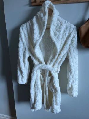 Vit fluffig morgonrock med huva - Mysig vit morgonrock med huva och knytskärp i midjan. Tillverkad i ett mjukt och fluffigt material som känns skönt mot huden. Perfekt för att chilla hemma och hålla sig varm. Långärmad och med avslappnad passform.