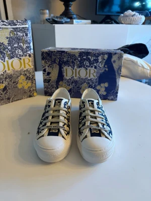 Dior skor - Snygga sneakers från Dior med vit bas och blått mönster samt logotypen 'Christian Dior' på tungan. Skorna har klassisk låg profil, vita snören och en stjärna på sulan. Perfekt för dig som vill sticka ut med exklusiva detaljer och trendig design.