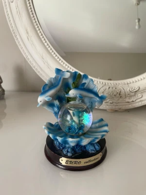 Dekorativ delfinprydnad med snöglob - Säljer en dekorativ prydnad från EURO collection med två blåvita delfiner, snäckskal och en snöglob i mitten. Prydnaden har havstema och är gjord i målad resin med detaljer i blått, vitt och grönt. Perfekt som inredningsdetalj för dig som gillar havet.
