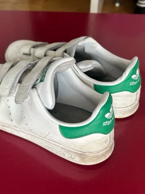 Adidas Stan Smith vita sneakers med kardborre - Klassiska vita Adidas Stan Smith sneakers med gröna detaljer på hälen och tungan. Skorna har tre kardborreband istället för snörning och är tillverkade i läder med en låg profil. Perfekt för dig som gillar retrostil och enkel design.