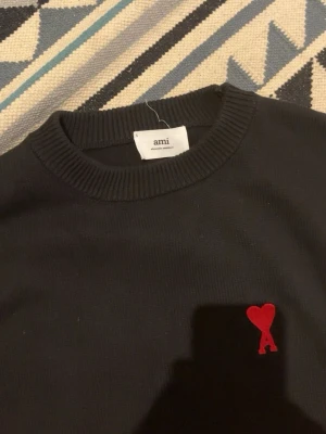 Svart tröja med röd Ami-logo - Cool svart tröja från Ami med deras ikoniska röda hjärt-logo broderad på bröstet. Tröjan har rund halsringning och ribbade muddar vid ärmslut och nederkant. Perfekt för dig som gillar stilrena plagg med en twist.
