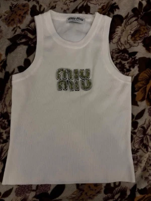 Vit ribbad tanktop från Miu Miu - Säljer en vit ribbad tanktop från Miu Miu med broderad logotyp framtill i grönt och silver. Toppen är ärmlös med rund halsringning och tillverkad i ett mjukt, stretchigt material. Perfekt för att skapa en trendig och avslappnad look.