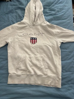 Vit hoodie från GANT  - Säljer en vit hoodie från GANT. Hoodien har stor huva, magficka och ribbade muddar. Tillverkad i mjukt bomullsmaterial, perfekt för en avslappnad och stilren look.