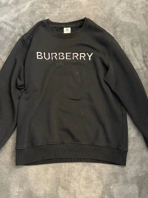 Svart Burberry sweatshirt med logo - Cool svart sweatshirt från Burberry med broderad logotyp och texten 'London England' på bröstet. Tröjan har rund halsringning, ribbade muddar och är tillverkad i mjukt material. Perfekt för dig som gillar stilrena designer och ikoniska märken.