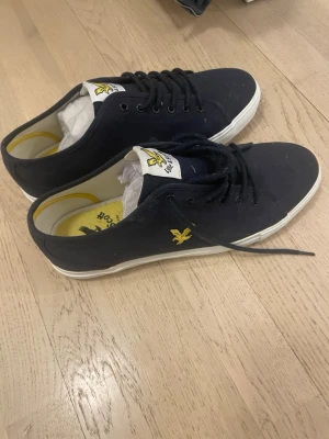 Blå Lyle & Scott sneakers - Snygga svarta sneakers från Lyle & Scott med klassisk låg profil. Skorna har vita sulor, svarta skosnören och detaljer i gult med logga på sidan och plös. Insidan är gul med tryckt logga. Perfekta för en stilren och avslappnad look. Helt nya med box å prislapp 