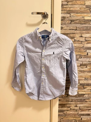 Ljusblå skjorta från Ralph Lauren - Klassisk ljusblå skjorta från Ralph Lauren med button-down krage och broderad logga på bröstet. Skjortan har långa ärmar, vita knappar och är tillverkad i bomull. Perfekt för en stilren och avslappnad look.