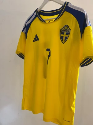 Svensk landslagströja Adidas gul och blå bergvall #7 - Gul svensk landslagströja från Adidas med blå detaljer, nummer 7 och namnet 'Bergvall' på ryggen. Tröjan har korta ärmar, broderad landslagssköld och Climacool-material som andas. Perfekt för att bära på matchdag och för en stundande vm💛💙