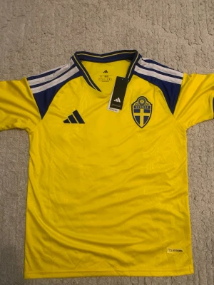 Svensk landslagströja Adidas gul - Gul svensk landslagströja från Adidas med blå detaljer och broderat landslagsemblem på bröstet. Tröjan har korta ärmar, blåa ränder på axlarna och är tillverkad i lätt och ventilerande material (Climacool). Perfekt för fotboll eller träning.