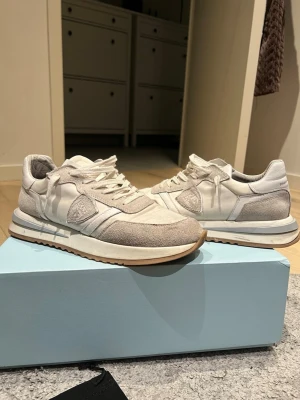 Vita Philippe Model sneakers - Säljer ett par snygga vita sneakers från Philippe Model med beige mockadetaljer och klassisk logga på sidan. Skorna har vita snören, gummisula och en stilren retroinspirerad design. Perfekta för dig som gillar minimalistisk och trendig stil. Skorna är köpte från merchsweden för ungefär 2750kr. Skorna är väll använda och sulorna har lossnat lite men det går att limma och man vill.