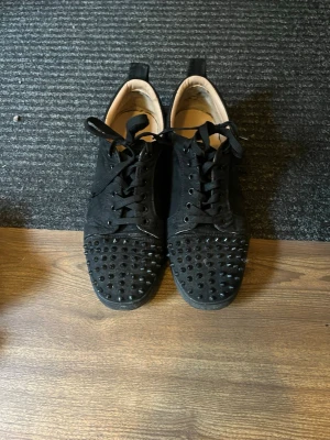 Louboutin skor - Säljer ett par svarta sneakers från Christian Louboutin med coola svarta nitar på tån och klassisk röd sula. Skorna har snörning, är låga och tillverkade i mocka och läder. Perfekta för dig som vill sticka ut med en edgy och lyxig stil.