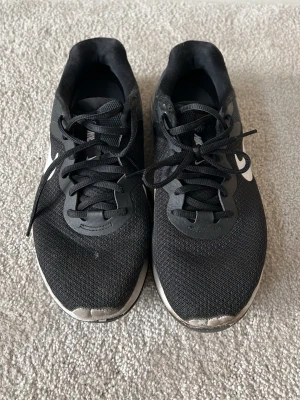 Svarta Nike sneakers med vit sula - Snygga svarta Nike sneakers med vit sula och klassisk swoosh-logga på sidan. Ovandelen är i meshmaterial för bra andningsförmåga och skorna har svarta skosnören. Yttersulan har ett mönstrat grepp för extra stabilitet.