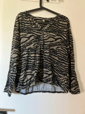 Zebramönstrad långärmad topp  - Cool långärmad topp med zebramönster i svart och beige. Toppen har v-ringning och är tillverkad i mjuk bomullsmix. Perfekt för dig som vill sticka ut med ett djurmönster i garderoben.