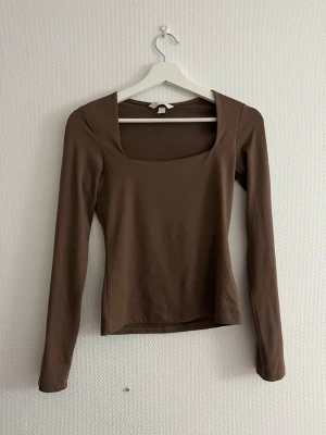 Brun långärmad topp med fyrkantig ringning - Snygg brun långärmad topp med fyrkantig ringning och figurnära passform. Perfekt att styla med jeans eller kjol för en clean look. Tillverkad i mjukt material som känns skönt mot huden.