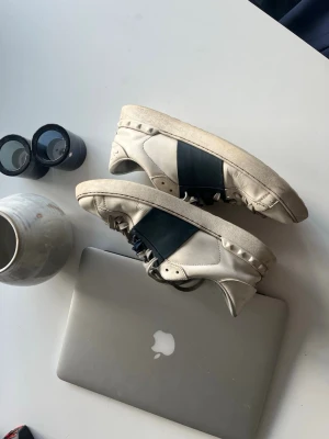 Vita Valentino sneakers med marinblå detalj - Snygga vita sneakers från Valentino med en bred svart läderdetalj över sidan. Skorna har lågt skaft, klassisk snörning och coola nitar längs sulan. Perfekta för dig som gillar stilrena men ändå unika sneakers.