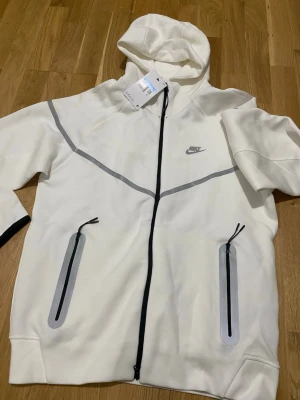 Vit Nike hoodie med dragkedja - Snygg vit hoodie från Nike med hel dragkedja framtill och två fickor med svarta dragkedjor. Hoodien har gråa detaljer över bröstet och en klassisk Nike-logga på vänster sida. Tillverkad i mjukt material och har en stor huva för extra komfort.