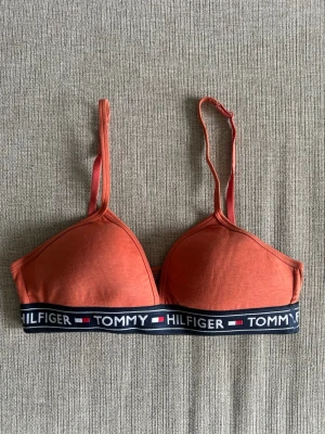 Orange bh från Tommy Hilfiger - Snygg orange bh från Tommy Hilfiger med justerbara axelband och elastiskt band med logga i marinblå, vitt och rött. OBS bh:n har ett litet hål på sidan (se bild 2)