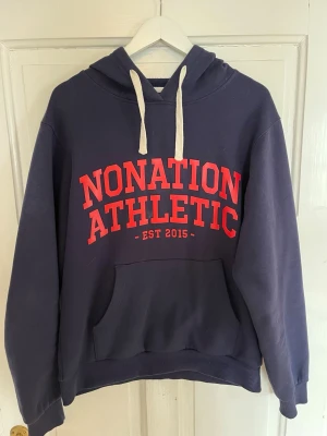Mörkblå hoodie Nonation Athletic - Mörkblå hoodie från Nonation Athletic med röd text och vit dragsko. Klassisk känguruficka framtill och ribbade muddar. Perfekt för en sportig och avslappnad stil. Tryck på bröstet: 'NONATION ATHLETIC EST 2015'.