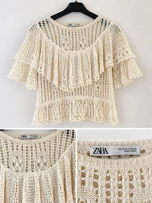 Krämvit virkad blus från Zara - Superfin krämvit virkad blus från Zara med bred volang över axlarna och ett luftigt, genombrutet mönster. Blusen har rund halsringning och korta ärmar. Perfekt för en trendig och bohemisk look.