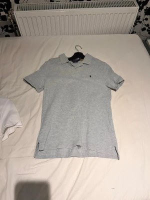 Grå pikétröja från Polo Ralph Lauren - Klassisk grå pikétröja från Polo Ralph Lauren med blå logga broderad på bröstet. Slim fit-modell med krage, knappar framtill och korta ärmar. Perfekt för en clean och stilren look.
