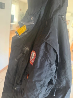 Svart parkasjacka från Parajumpers - Säljer en svart parkasjacka från Parajumpers med ikonisk patch på ärmen och gul detalj vid kragen. Jackan har flera fickor med knappar och dragkedjor samt robusta sömmar. Perfekt för kalla dagar och riktigt snygg streetstil.