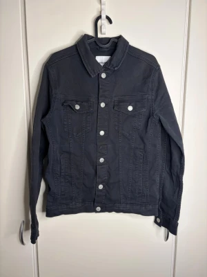 Svart jeansjacka från Jack & Jones - Stilren svart jeansjacka från Jack & Jones med klassisk krage, silverfärgade knappar och två bröstfickor med lock. Jackan har raka linjer och är tillverkad i slitstarkt jeanstyg. Perfekt för en cool och avslappnad look.