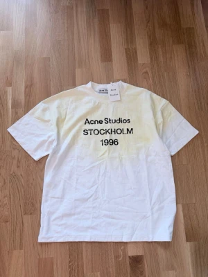 Vit Acne Studios t-shirt med tryck - Vit t-shirt från Acne Studios med svart texttryck 'Acne Studios STOCKHOLM 1996' på bröstet. Klassisk passform med rund hals och korta ärmar. Perfekt för en stilren och trendig look.