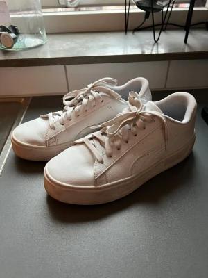 Vita Puma sneakers i läder - Snygga vita sneakers från Puma med klassisk låg siluett. Skorna är gjorda i fejkläder med Pumas ikoniska formstripe på sidan och logga på sulan. Perfekta för en clean och stilren look. Skosnören och sula i vitt för en enhetlig vibe. Endast använda ett par gånger. Mycket bra skick!