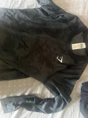 Svart långärmad träningströja Gymshark - Svart långärmad träningströja från Gymshark med diskret camomönster och vitt logotryck på bröstet. Tillverkad i ett lätt och stretchigt funktionsmaterial som andas, perfekt för gym eller löpning.