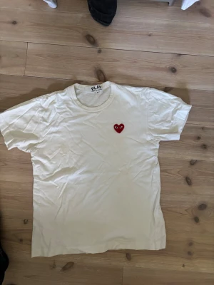 Vit t-shirt från Comme des Garçons Play - Vit t-shirt från Comme des Garçons Play med det ikoniska röda hjärtat med ögon broderat på bröstet. Klassisk rund hals och korta ärmar. Perfekt för en stilren och trendig look.