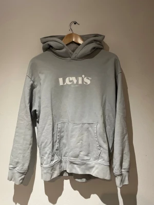 Levi's Hoodie - Snygg grå/blå hoodie från Levi's med stor vit logga på bröstet. Klassisk modell med huva och känguruficka framtill. Tillverkad i mjukt bomullsmaterial och har ribbade muddar vid ärmslut och nederkant. Perfekt för en avslappnad och trendig stil.