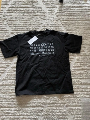 Maison Margiela svart t-shirt - Svart t-shirt från Maison Margiela med ikoniskt tryck framtill i vitt med siffror och logga. Klassisk passform med rund hals och korta ärmar. Perfekt för dig som gillar stilrena och trendiga plagg med designer-touch.