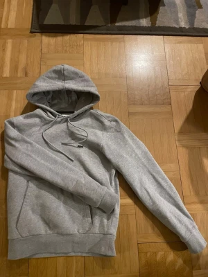 Grå hoodie från Lacoste - Snygg grå hoodie från Lacoste med klassisk krokodillogga på bröstet. Hoodien har dragsko i huvan, magficka och ribbade muddar vid ärmslut och nederkant. Perfekt för en avslappnad och stilren look.