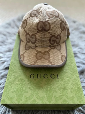 Beige keps med GG-mönster - Snygg beige keps med klassiskt GG-monogram i brunt över hela kepsen. Kepsen har böjd skärm, justerbar rem bak och är tillverkad i canvasmaterial. Perfekt för dig som vill ha en trendig accessoar med lyxig känsla.