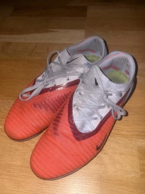Nike röda och vita fotbollsskor - Säljer ett par Nike fotbollsskor i rött och vitt med svarta detaljer. Skorna har snörning, låg profil och mönstrad ovandel för bra bollkontroll. Yttersulan har fasta dobbar som passar för spel på gräs. Snygg och sportig design som sticker ut på planen.