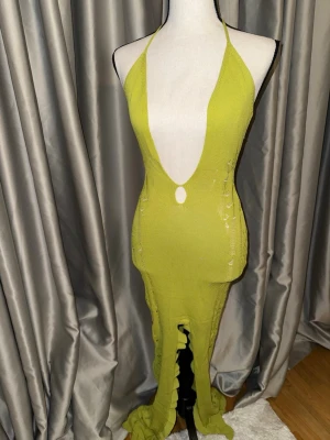 Murci Lång limegrön stickad klänning - Långklänning i limegrönt stickat material med djup v-ringning och halterneck. Klänningen har en öppen rygg och dekorativa detaljer längs sidorna samt volangkant längst ner. 