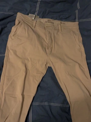 Beige chinos från Levi's - Snygga beige chinos från Levi's med klassisk passform och raka ben. Byxorna har sidofickor fram och en bakficka med knapp samt Levi's logotyp på baksidan. Perfekta för dig som vill ha ett stilrent och bekvämt plagg.