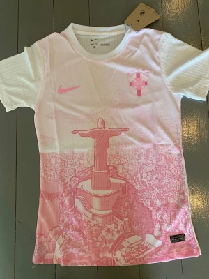 Vit och rosa Brasilien fotbollströja - Nike fotbollströja i vitt och rosa med CBF-logga och ikoniskt motiv av Kristusstatyn i Rio de Janeiro. Tröjan har rund hals och korta ärmar, tillverkad i ett lätt och ventilerande material. Perfekt för dig som älskar fotboll och vill sticka ut på planen.