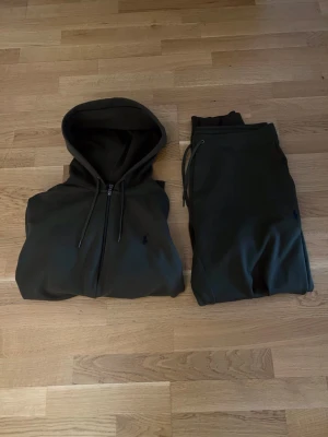 Polo Ralph lauren  - Snyggt mörkgrönt set från Polo Ralph Lauren med hoodie och matchande byxor. Hoodien har dragkedja och dragsko, byxorna har resår och snörning i midjan. Perfekt för en avslappnad och sportig look.