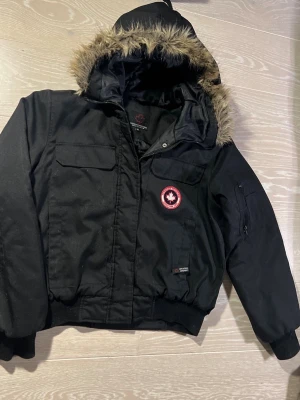 Svart parkas från Canadian Mountain XL - Svart parkasjacka från Canadian Mountain med fuskpäls på huvan och flera fickor framtill. Jackan har en röd logotyp på bröstet och muddar vid ärmslut och midja. Perfekt för kalla dagar med sin varma och robusta design.