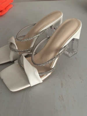 Vita pumps med transparent klack - Snygga vita pumps med fyrkantig tå, två glittriga remmar och en bredare tvinnad rem framtill. Klacken är transparent och chunky vilket ger en modern look. Perfekta för dig som vill sticka ut med stilrena detaljer.