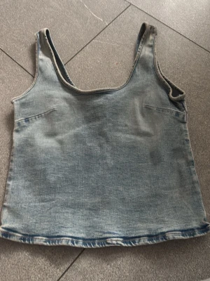 Ljusblå denim linnetopp - Säljer en ljusblå linnetopp i denim-look med breda axelband och rundad halsringning. Toppen har en croppad passform och är perfekt för dig som gillar en avslappnad och trendig stil. Inga synliga märken eller loggor.