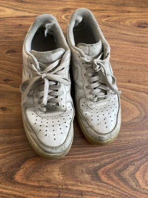 Vita Nike Air Force 1 sneakers - Klassiska vita Nike Air Force 1 sneakers i läder med perforerad tå, vita skosnören och den ikoniska swoosh-loggan på sidan. Yttersulan är något gulnad och skon har en låg silhuett. Perfekta för dig som gillar streetwear och tidlös design.