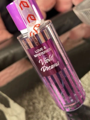 Love & Mysteries Violet Dreams Bodymist - Snygg bodymist i lila och genomskinlig plastflaska med glittriga detaljer och randigt mönster. Doften heter Violet Dreams och flaskan har ett matchande lila lock. Perfekt för dig som gillar fräscha och blommiga dofter.